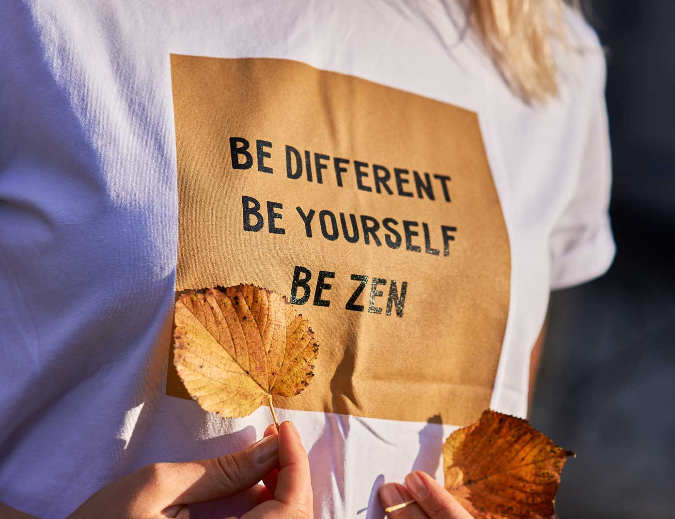 Ein weißes T-Shirt mit der inspirierenden Aufschrift „BE DIFFERENT BE YOURSELF BE ZEN“ wird in natürlicher Umgebung mit Herbstblättern präsentiert – Sinnstoff