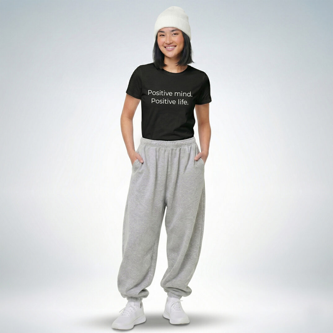 Weibliches Model mit weißer Mütze trägt schwarzes T-Shirt mit „Positive mind. Positive life.“ Print und graue Jogginghose – Sinnstoff