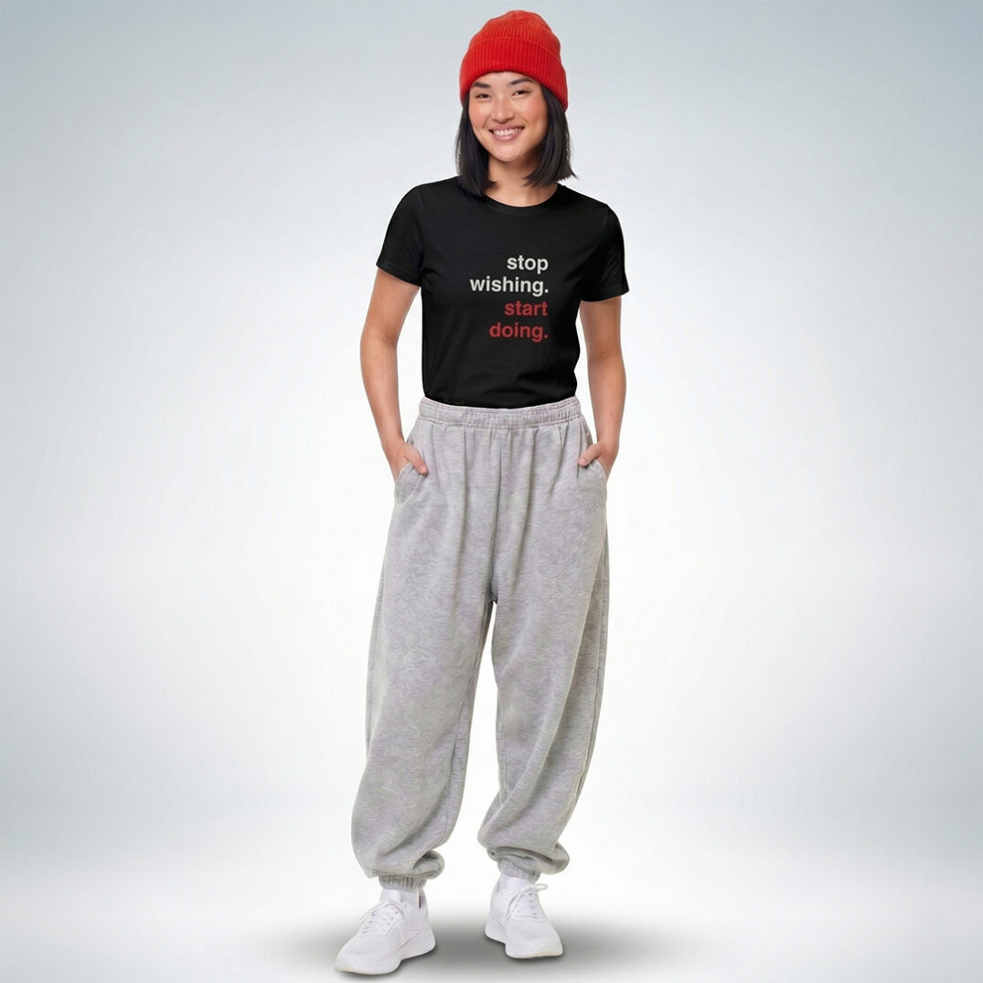 Weibliches Model mit roter Mütze trägt schwarzes T-Shirt mit „Stop wishing. Start doing.“ Print und graue Jogginghose – Sinnstoff
